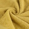 Musztardowy ręcznik o fakturze frotte, z widocznym wzorem geometrycznym na brzegu, ułożony w miękkie fałdy. Materiał wygląda na puszysty i miękki w dotyku.