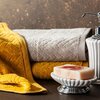 Na zdjęciu widoczny jest musztardowy ręcznik o wzorze w jodełkę, złożony obok szarego ręcznika, a obok nich znajduje się biała mydelniczka z mydłem oraz dozownik na mydło w płynie. Tło jest ciemne, z delikatnymi złotymi akcentami.