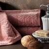 Pudrowy ręcznik o wymiarach 70 x 140 cm z widocznym wzorem w jodełkę leży obok białego dozownika na mydło i mydelniczki z beżowym mydłem. Na pierwszym planie znajdują się dwa dekoracyjne orzechy.