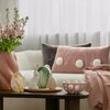 Na zdjęciu znajduje się jasna sofa z dwoma poduszkami, jedna z nich to poszewka EUROFIRANY 467106 Wiosna 54 30 x 50 cm Pudrowy, ozdobiona białymi pomponami. Przed sofą stoi stolik z dekoracyjnymi jajkami, figurką królika i różową wazonem z kwiatami.