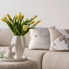 Na zdjęciu znajduje się jasna sofa z dwoma poduszkami ozdobionymi motywem zająca oraz białym wazonem z żółtymi i białymi tulipanami. Obok wazonu stoi figurka zająca na okrągłej tacy.