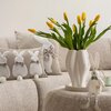 Na zdjęciu znajduje się jasna sofa z beżowymi poduszkami ozdobionymi motywem królików oraz białym wazonem z żółtymi tulipanami. Obok wazonu stoi pluszowy królik, a całość tworzy wiosenny wystrój wnętrza.