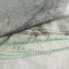 Zdjęcie przedstawia fragment pościeli w jasnoszarym kolorze z delikatnym wzorem w odcieniach zieleni i szarości. Na materiale widoczne jest logo oraz napis "EVA MINGE".