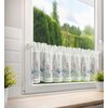 Biała firana o wymiarach 150 x 30 cm wisi na oknie, ozdobiona jest delikatnym wzorem kwiatowym w odcieniach różu i niebieskiego oraz koronkowym wykończeniem na dole. W tle za oknem widoczny jest rozmyty krajobraz z zielonymi drzewami i niebem.
