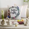 Na zdjęciu znajduje się biała poszewka z napisem "happy Easter" otoczonym kwiatowym wieńcem i wizerunkiem królika. W tle widoczna jest druga poszewka, a na pierwszym planie dekoracje wielkanocne, w tym ceramiczne figurki królików, miska z kolorowymi jajka