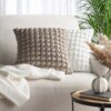 Na zdjęciu znajduje się jasna sofa z dwoma poduszkami o teksturowanej powierzchni, jedna w kolorze beżowym, druga biała. Obok sofy stoi okrągły stolik z metalowymi nogami, na którym leży książka i stoi wazon z suszonymi trawami.