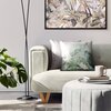 Na zdjęciu znajduje się jasna sofa z poduszką w zielone liście, obok której stoi lampa podłogowa i roślina doniczkowa. Przed sofą znajduje się okrągły puf z książkami i kubkiem, a na ścianie wisi obraz z motywem roślinnym.