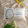 Na zdjęciu znajduje się biała poszewka z napisem "Wishing you a Happy Easter" oraz grafiką królika i kwiatów, leżąca na beżowym materiale. Wokół poszewki widoczne są żółte i białe tulipany.