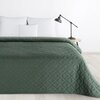 Ciemnozielona narzuta o wymiarach 170 x 210 cm z geometrycznym wzorem w romby, rozłożona na łóżku z białymi poduszkami. Obok łóżka znajduje się czarna lampa na drewnianym stoliku.