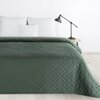 Zielona narzuta o wymiarach 220 x 240 cm z geometrycznym wzorem leży na łóżku z białymi poduszkami, obok znajduje się czarna lampa na drewnianym stoliku. Ściana w tle jest biała, a podłoga ma jasny, drewniany kolor.