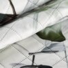 Zbliżenie na białą narzutę z pikowanym wzorem w romby, ozdobioną motywem roślinnym w odcieniach zieleni i szarości. Tkanina ma delikatną teksturę i widoczne przeszycia.