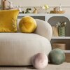 Na zdjęciu znajduje się jasna sofa z musztardową poduszką i okrągłą poduszką w tym samym kolorze. W tle widoczny jest biały regał z dekoracjami, takimi jak figurki królików, książki i rośliny.
