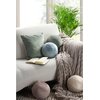 Na zdjęciu znajduje się jasna sofa z kilkoma poduszkami w różnych odcieniach zieleni i beżu oraz otwarta książka. Obok sofy leży miękki, szary koc, a w tle widoczna jest zielona roślina doniczkowa.