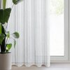 Na zdjęciu znajduje się jasna firana zawieszona na karniszu, przez którą widać zewnętrzny krajobraz z drzewami. Przed oknem stoi jasnoniebieska sofa z kilkoma poduszkami, a obok niej znajduje się drewniany stolik z dekoracyjnym przedmiotem.