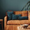 Na zdjęciu znajduje się brązowa, skórzana sofa z trzema poduszkami w niebieskich poszewkach o różnym kształcie. Obok sofy stoi drewniany stolik z metalową misą i kadzidłem, a w tle widać zieloną ścianę oraz gałązki rośliny.