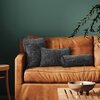 Na zdjęciu znajduje się brązowa, skórzana sofa, na której leżą trzy poduszki w odcieniach szarości. W tle widoczna jest ciemnozielona ściana, a na pierwszym planie drewniany stolik z metalową misą i kadzidłem.