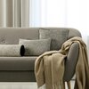 Na zdjęciu znajduje się elegancka sofa w odcieniu beżu, na której ułożone są trzy poduszki w różnych kształtach i rozmiarach, wszystkie w stonowanych kolorach z ciemnymi obwódkami. Na oparciu fotela obok sofy leży beżowy koc z frędzlami, a w tle widoczne 