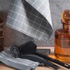 Na zdjęciu widoczne są dwie srebrne ściereczki z wzorem w kratkę, jedna zawieszona, a druga złożona na blacie. Obok nich znajdują się czarne przybory kuchenne oraz szklany pojemnik w kolorze bursztynowym.