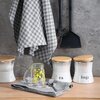 Na zdjęciu widoczna jest szara ściereczka w kratkę zawieszona na ścianie, obok której znajdują się dwa czarne przybory kuchenne. Na blacie stoją trzy białe pojemniki z drewnianymi pokrywkami, z napisami "Tea" i "Sugar", a obok nich leży szklanka z żółtym 