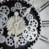 Zegar ścienny z widocznymi zębatkami w kolorach srebrnym i czarnym, z białymi wskazówkami na tle tarczy z rzymskimi cyframi. Tło zegara jest w odcieniach bieli i szarości z efektem postarzenia.
