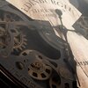 Zbliżenie na mechanizm zegara ściennego z widocznymi zębatkami i wskazówkami na tle drewnianej tarczy. Na tarczy widoczny jest tekst: "EDINBURGH CLOCK WORKS COMPANY EST. 1923".