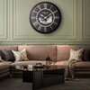 Na zdjęciu znajduje się elegancki salon z dużym, czarno-srebrnym zegarem ściennym z rzymskimi cyframi i napisem "Oldgate". Wnętrze uzupełnia brązowa sofa z poduszkami oraz szklany stolik z czarną tacą, na której stoi dzbanek i filiżanka.