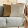 Na zdjęciu widoczna jest szara sofa z dwiema poduszkami: ciemnobeżową i brązową, oraz beżowym kocem z frędzlami. W tle znajduje się drewniana żaluzja.