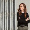 Na zdjęciu widoczna jest zasłona w kolorze srebrno-złotym z wzorem przypominającym liście lub pióra. Obok zasłony stoi osoba ubrana w ciemny sweter i spodnie, z założonymi rękami.