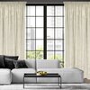 Na zdjęciu znajduje się jasna zasłona o wymiarach 140 x 270 cm w kolorze kremowym, zawieszona przy dużym oknie z widokiem na miasto. Przed oknem stoi jasna sofa z ciemnymi poduszkami oraz czarny stolik z misą pełną zielonych jabłek.