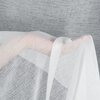 Biała firana o delikatnej, przezroczystej strukturze jest ułożona na tle szarej, teksturowanej powierzchni. Materiał firany ma subtelny wzór, który dodaje elegancji i lekkości.