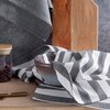 Na zdjęciu widoczna jest ściereczka w szaro-białe pasy, ułożona obok szklanej miski i drewnianej deski. W tle znajduje się słoik z drewnianą pokrywką, wypełniony ciemnymi ziarnami.