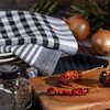 Na zdjęciu widoczne są trzy ściereczki w czarno-białą kratkę i paski, ułożone na stosie talerzy. Obok znajdują się suszone papryczki na drewnianej desce, słoik z przyprawami oraz cebule w tle.