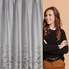 Srebrna zasłona EUROFIRANY Mariela o wymiarach 140 x 270 cm z subtelnym wzorem na dolnej części. Obok zasłony widoczna jest osoba w czarnym swetrze i brązowych spodniach na beżowym tle.