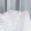 Na zdjęciu widoczna jest biała firana z delikatnego, przezroczystego materiału, ozdobiona falbanami. W tle znajduje się gładka, szara zasłona.
