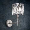 Kinkiet z metalowym ramieniem i okrągłą podstawą, posiada klosz z przezroczystych, kryształowych elementów, zamontowany na tle ciemnej, teksturowanej ściany. Światło lampy odbija się w kryształach, tworząc efektowne refleksy.