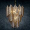Lampa sufitowa o kształcie przypominającym liście, wykonana z materiału w kolorze grafitowo-złotym, emituje ciepłe światło. Tło jest ciemne, co podkreśla detale i strukturę lampy.