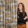 Zasłona w kolorach musztardowym i srebrnym z abstrakcyjnym wzorem, obok której stoi osoba w czarnym swetrze i brązowych spodniach na tle beżowej ściany. Osoba ma długie, falowane włosy i uśmiecha się, patrząc w bok.