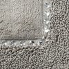 Srebrny dywanik łazienkowy o wymiarach 50 x 70 cm leży na drewnianej podłodze. W prawym górnym rogu znajduje się powiększenie pokazujące teksturę i detale materiału.