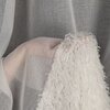 Na zdjęciu widoczna jest biała firana o teksturze przypominającej delikatne pióra, zawieszona na tle szarej, półprzezroczystej tkaniny. Produkt to "Firana gotowa EUROFIRANY Marion 140 x 250 cm Biały".