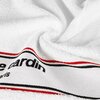 Biały ręcznik z widocznym czarnym napisem "Pierre Cardin Paris" na białym pasku z czerwonymi i czarnymi liniami. Materiał ręcznika jest puszysty i miękki, z lekkimi zagnieceniami.
