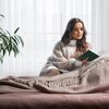 Na zdjęciu widoczny jest różowy koc z frędzlami, rozłożony na łóżku, obok którego znajduje się roślina doniczkowa. W tle widać jasne zasłony przepuszczające światło.