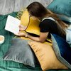 Na zdjęciu widać osobę leżącą na brzuchu, czytającą książkę, otoczoną różnokolorowymi poduszkami, w tym ciemnoniebieską poszewką EUROFIRANY Mel 2 o wymiarach 40 x 40 cm. Na podłodze leży zielony koc, a obok znajduje się filiżanka z napojem.