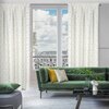 Na zdjęciu znajduje się elegancki salon z dużymi oknami, przez które widać balkon z ozdobną balustradą i panoramę miasta. W pomieszczeniu znajduje się zielona sofa z poduszkami, szklany stolik z dekoracjami oraz białe zasłony EUROFIRANY Marigold.
