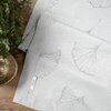 Na zdjęciu widoczny jest szary obrus z delikatnym wzorem liści, z metką z napisem "Eurofirany". Obrus leży na drewnianym stole obok zielonej rośliny i metalowej miski.