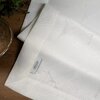 Biały bieżnik z delikatnym wzorem leży na drewnianym stole, obok znajduje się roślina i miska. Na bieżniku widoczna jest metka z napisem "Eurofirany".