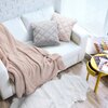 Na zdjęciu znajduje się jasna sofa z dwoma poduszkami o pikowanym wzorze, jedna w kolorze jasnoróżowym, a druga szara, oraz jasnoróżowy koc. Obok sofy stoi żółty stolik z magazynami, a na podłodze leży biały, puszysty dywan.