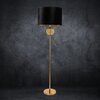 Czarna lampa podłogowa z cylindrycznym abażurem i złotą podstawą, ozdobiona przezroczystą kulą na środku trzonu, stoi na tle ciemnej, teksturowanej ściany. Produkt to "Lampa podłogowa EUROFIRANY Leona 467319 Czarno-złoty wysokość 165 cm".