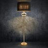 Na zdjęciu widoczna jest lampa stojąca z czarnym abażurem i złotą podstawą, umieszczona na tle ciemnej, teksturowanej ściany. Lampa ma kulisty element dekoracyjny pod abażurem, który odbija światło.