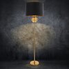 Czarna lampa podłogowa z okrągłym, złotym elementem na środku i złotą podstawą stoi na tle ciemnej, teksturowanej ściany. Abażur jest cylindryczny, a światło tworzy delikatny wzór na ścianie.