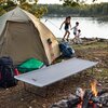Na zdjęciu znajduje się szare łóżko turystyczne ustawione na trawie obok namiotu, z widocznym logo "NILS CAMP". W tle widać jezioro oraz ognisko, a obok namiotu leżą plecaki i kijki trekkingowe.